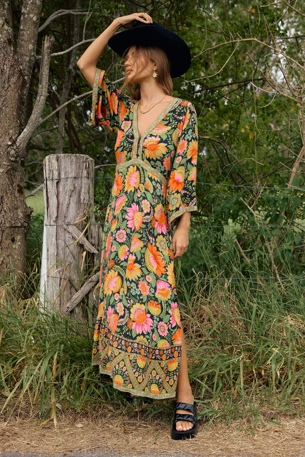 Woman in a colorful floral dress standing outdoors near a wooden fence.