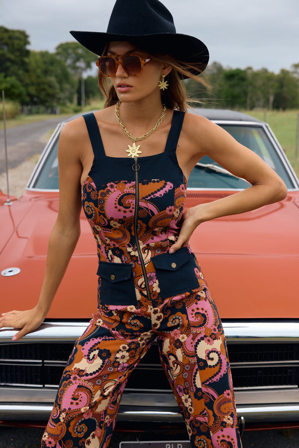 Woman in a patterned jumpsuit and black hat standing in front of a vintage car.
