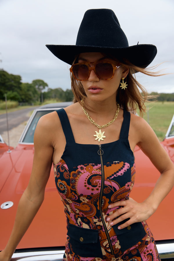 Woman wearing a black hat and colorful top with sunglasses, standing in front of a red car.