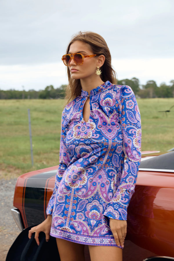 Woman in a blue and pink patterned dress standing next to a vintage car in an open field.