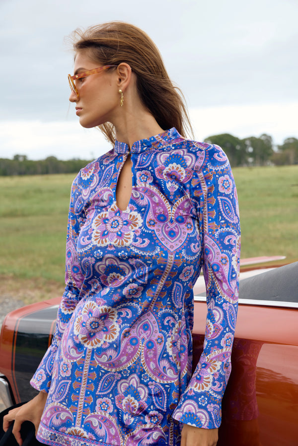 Woman wearing a blue and purple patterned dress standing next to a red car with a field in the background.