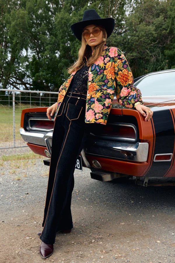 Person wearing a floral jacket and black hat leaning against a vintage car with trees in the background