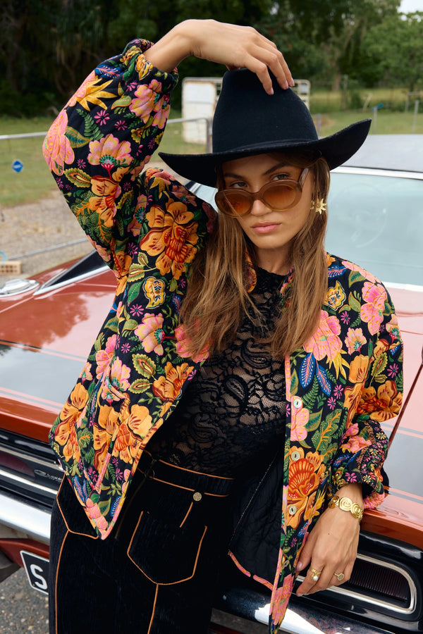 Woman wearing a colorful floral jacket and black hat in front of a vintage car.