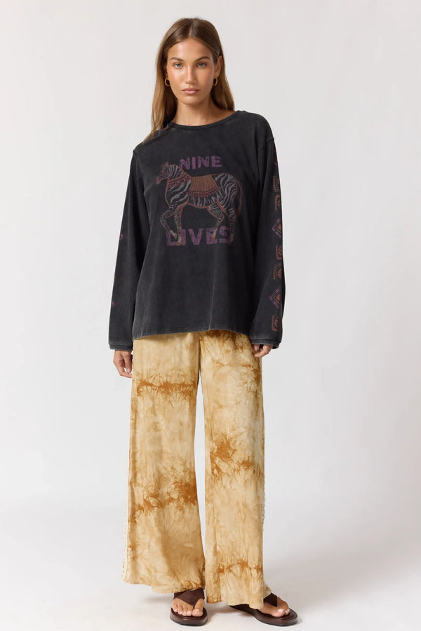 Woman wearing a black long-sleeve shirt with a graphic design and beige tie-dye pants on a white background