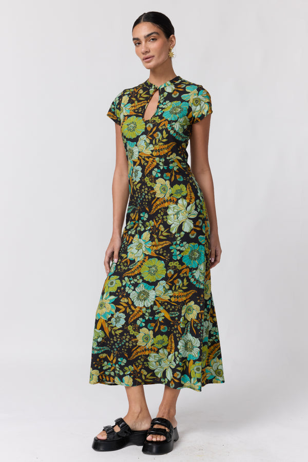 Nine Lives Bazaar Nostalgia Willa Maxi Dress. A woman modeling a short-sleeve midi dress with a vibrant floral print in green, yellow, and blue on a dark background, paired with black platform sandals, against a light gray studio backdrop.
