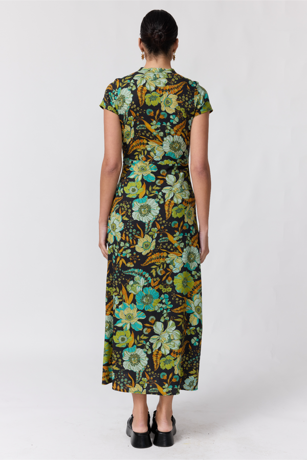 Nine Lives Bazaar Nostalgia Willa Maxi Dress. A back shot of a woman modeling a short-sleeve midi dress with a vibrant floral print in green, yellow, and blue on a dark background, paired with black platform sandals, against a light gray studio backdrop.
