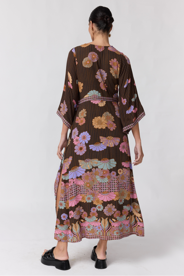 A woman viewed from the back wearing a brown kimono-style maxi dress with a colorful floral and butterfly print, paired with black slip-on shoes, against a light gray studio background.