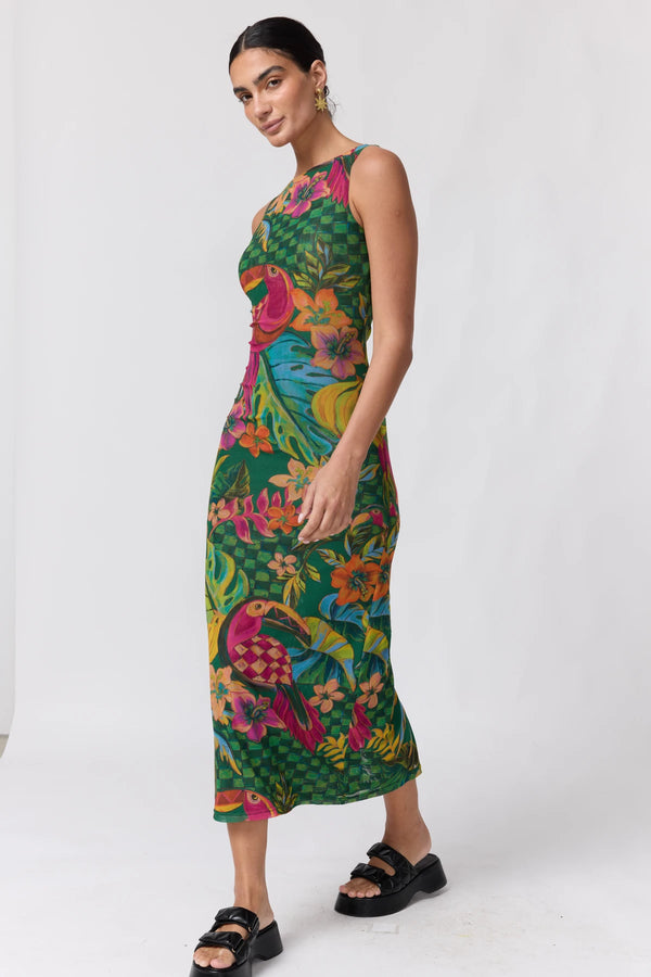 A woman modeling a sleeveless, high-neck midi dress with a vibrant tropical print featuring green foliage and colorful birds, paired with black slide sandals against a plain white studio background.