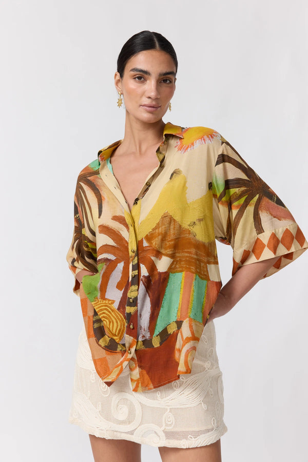 A woman modeling a short-sleeve button-up shirt with a vibrant, multicolor abstract tropical print in orange, yellow, brown, and green, paired with a white lace skirt against a light gray studio background.