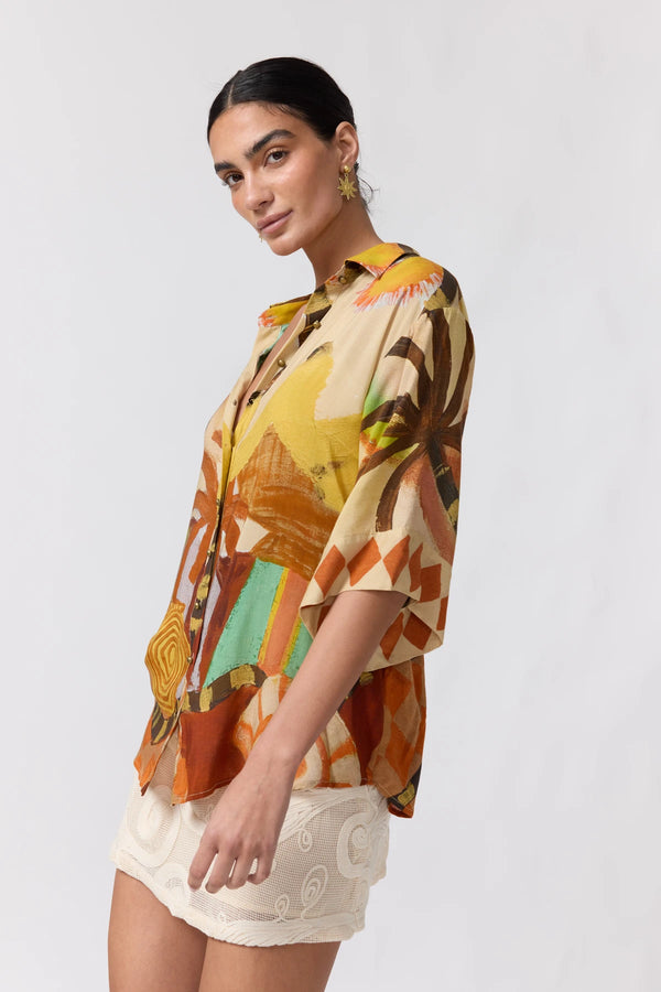 A woman modeling a short-sleeve button-up shirt with a colorful abstract print in orange, yellow, brown, and green, paired with cream lace shorts against a light gray studio background.
