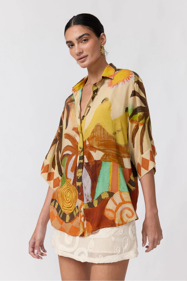 A woman modeling a short-sleeve button-up shirt with a vibrant abstract print in orange, yellow, brown, and green on a cream base, paired with a white textured skirt against a light gray studio background.