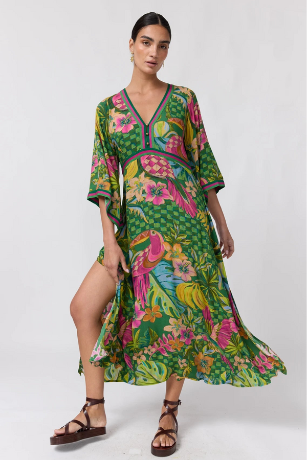 A woman modeling a vibrant green tropical print kaftan dress with pink flamingos, toucans, and colorful flowers, paired with brown strappy sandals against a plain white studio background.