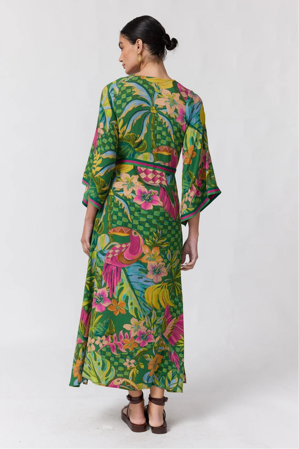 A woman shown from the back wearing a long-sleeved maxi dress with a vibrant tropical print featuring green palm trees, pink flamingos, and yellow birds on a light gray studio background.