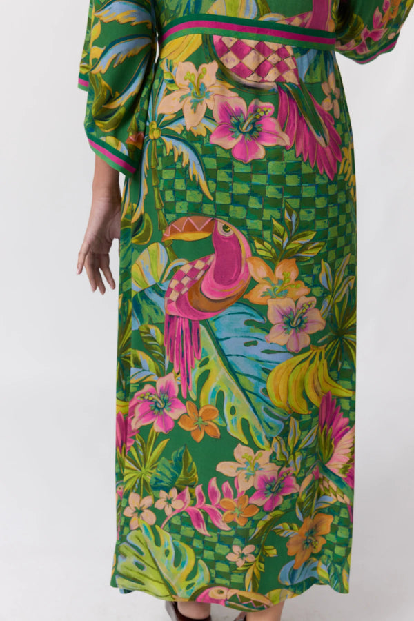 A close-up of a person wearing a vibrant green tropical print maxi dress with a toucan, flowers, and checkered patterns, shown against a plain white background.
