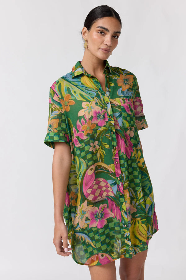 A woman modeling a short-sleeve button-up shirt dress with a vibrant green tropical print featuring pink flamingos, orange flowers, and a checkered pattern, set against a plain light gray background.