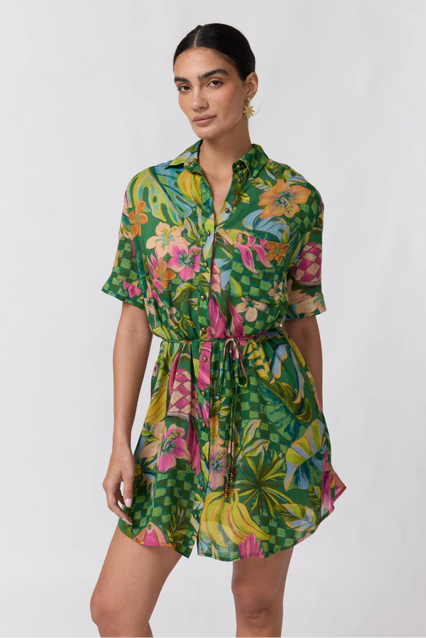 A woman modeling a short-sleeve collared romper dress with a vibrant green tropical floral print, featuring pink, yellow, and orange flowers, set against a plain light gray background.