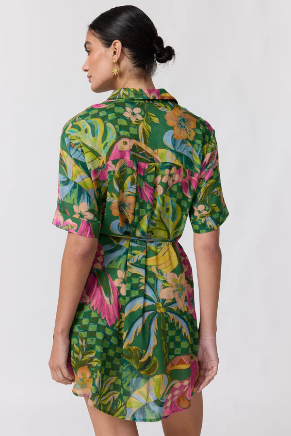 A woman shown from the back wearing a short-sleeve collared shirt dress with a vibrant tropical print featuring green, pink, yellow, and blue. The dress has a self-tie waist and is photographed against a plain light gray background.