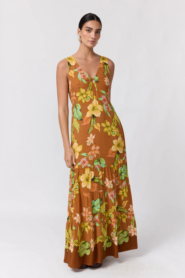 A woman modeling a sleeveless, tiered maxi dress with a brown base and a vibrant yellow, green, and orange floral print, set against a plain white studio background.
