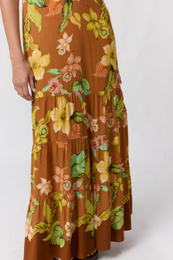 A close-up of a tiered maxi skirt with a brown base and a vibrant floral print featuring yellow, pink, and green flowers and leaves, shown on a model against a light gray background.