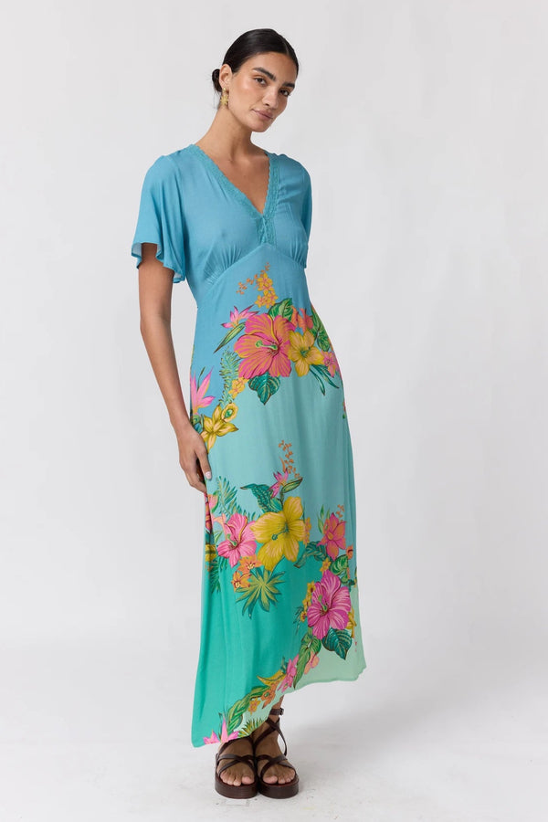 A woman modeling a turquoise maxi dress with a vibrant tropical floral print in pink, yellow, and green, styled with brown strappy sandals against a plain white studio background.