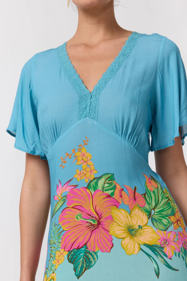 A woman wearing a light blue short-sleeve V-neck dress with a lace trim at the neckline and a colorful tropical floral print in pink, yellow, and green across the lower front.
