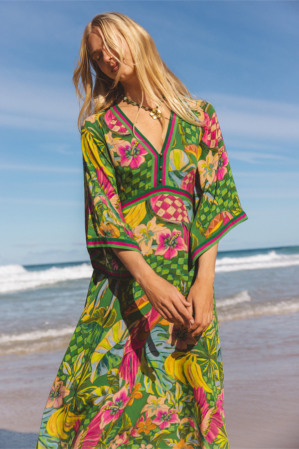 A woman wearing a vibrant green tropical print kaftan dress stands on a sandy beach with the ocean and blue sky in the background. Location is Dreamtime Beach, Fingal Head, NSW.