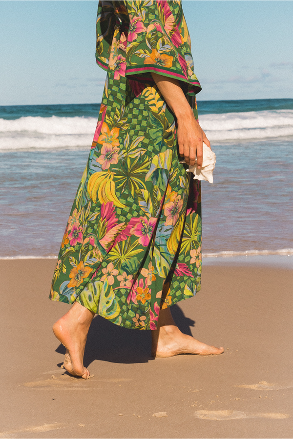 A person walking barefoot on a sandy beach wearing a long, wide-sleeved tropical floral dress or cover-up in green with pink, yellow, and orange floral patterns. The ocean and blue sky are visible in the background.