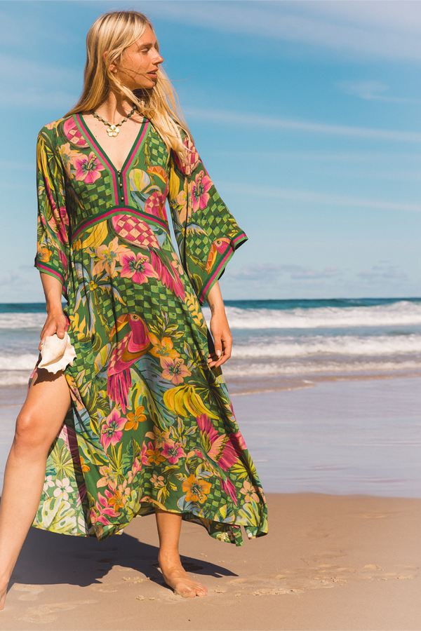 A woman stands on a sandy beach wearing a flowing tropical print maxi dress with wide sleeves. The dress has a green base with pink, yellow, and orange floral and bird motifs, and is photographed against a blue sky and ocean background.