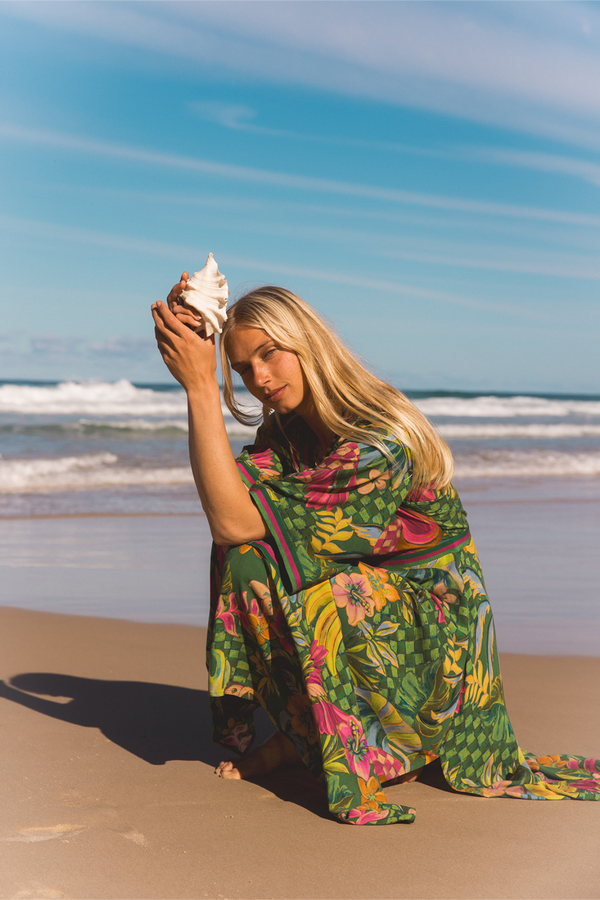 A woman kneeling on a sandy beach with the ocean and blue sky behind her, wearing a flowing green tropical floral print wrap or dress and holding a large white seashell. Location is Dreamtime Beach, Fingal Head, NSW.