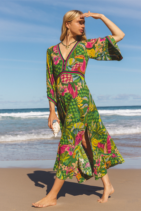 A woman stands barefoot on a sandy beach wearing a vibrant tropical print maxi dress with a wrap-style V-neck, wide sleeves, and a side slit. She holds a white shell against the blue ocean and sky background.