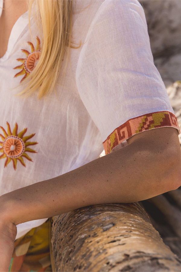 A close-up lifestyle shot of a woman wearing a white linen blouse with embroidered sun motifs and a patterned cuff, resting her arm on a tree trunk outdoors.