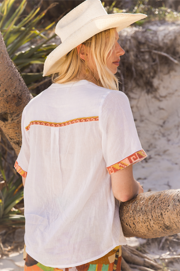 A woman wearing a white short-sleeve bohemian-style shirt with orange and red patterned trim on the back and sleeve cuffs, paired with a cream cowboy hat, photographed outdoors on a sandy beach with palm trees.