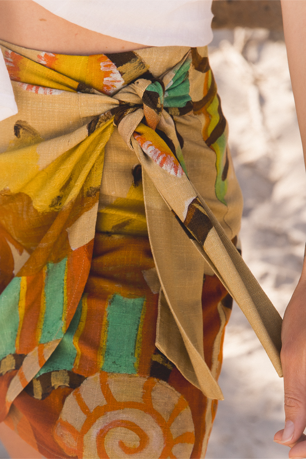 A close-up of a person wearing a colorful patterned sarong wrap tied around the waist, with a white top visible at the top edge. The wrap features a mix of mustard yellow, orange, beige, green, and brown abstract and geometric patterns.