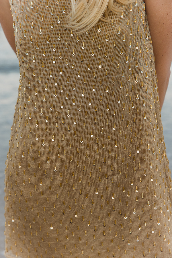 Close-up of a sleeveless textured top or dress in warm beige with an allover pattern of small gold and silver teardrop-shaped sequins, worn by a model with blonde hair visible at the top. 