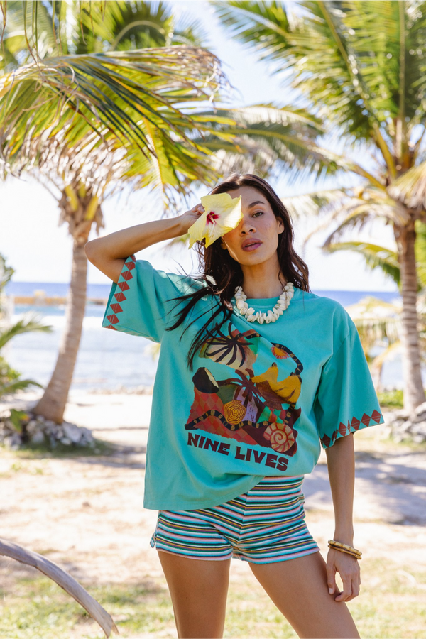 A woman posing on a tropical beach with a yellow hibiscus and wearing a turquoise graphic t-shirt with 'NINE LIVES' text and a colourful illustration, paired with striped shorts. Location is Cook Islands.