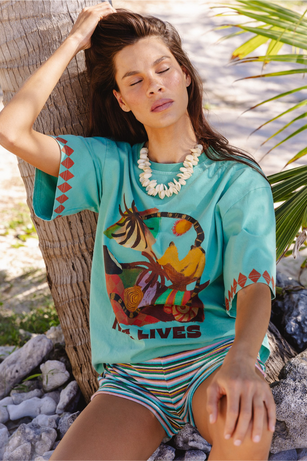 A woman reclines against a palm tree outdoors, wearing a turquoise graphic T-shirt with a multicolor animal print and striped shorts in a beach-like setting.