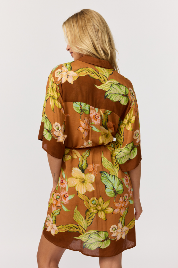 A woman shown from the back wearing a brown short-sleeve wrap dress with a vibrant green, yellow, and orange tropical floral print, set against a plain light gray background.