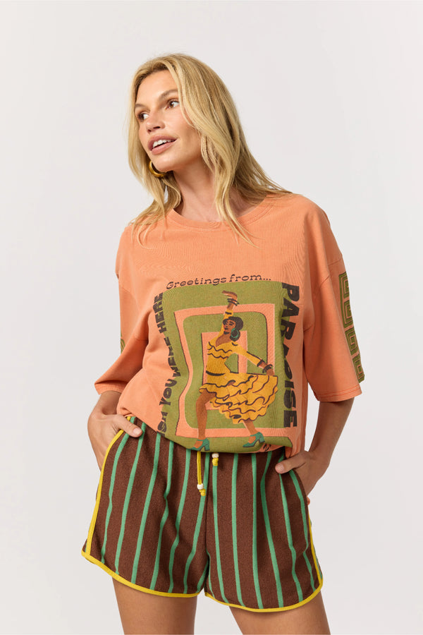 Woman wearing a peach-colored t-shirt with graphic design and brown shorts with green stripes on a white background