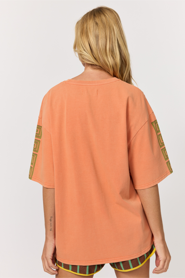 A woman viewed from the back wearing an oversized peach-colored t-shirt with olive green and yellow geometric patterns on the sleeves, paired with striped shorts.