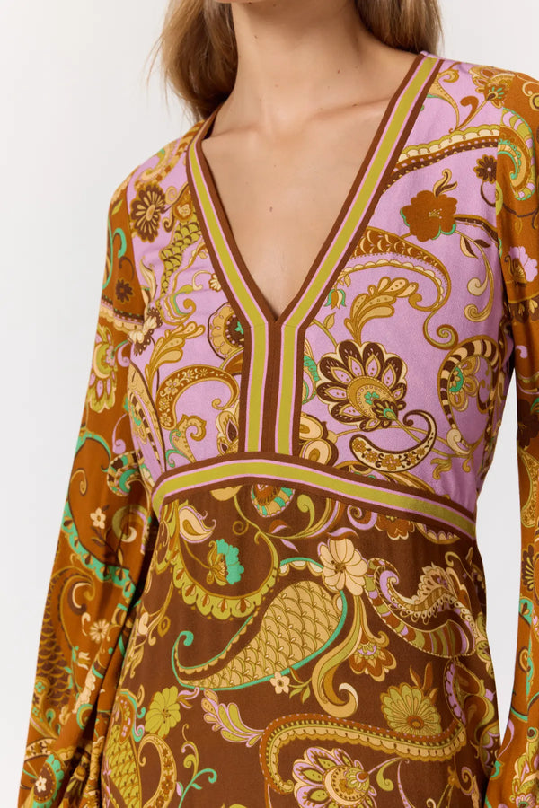 A close-up of a long-sleeve V-neck dress featuring a detailed paisley and floral print. The top portion is light lavender pink with gold and brown patterns, while the lower portion is a rich brown with gold and teal accents.