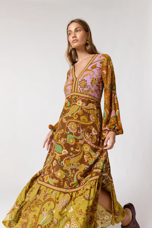 A woman modeling a long-sleeved maxi dress with a complex paisley and floral pattern in yellow, brown, and lavender, set against a plain light gray studio background.