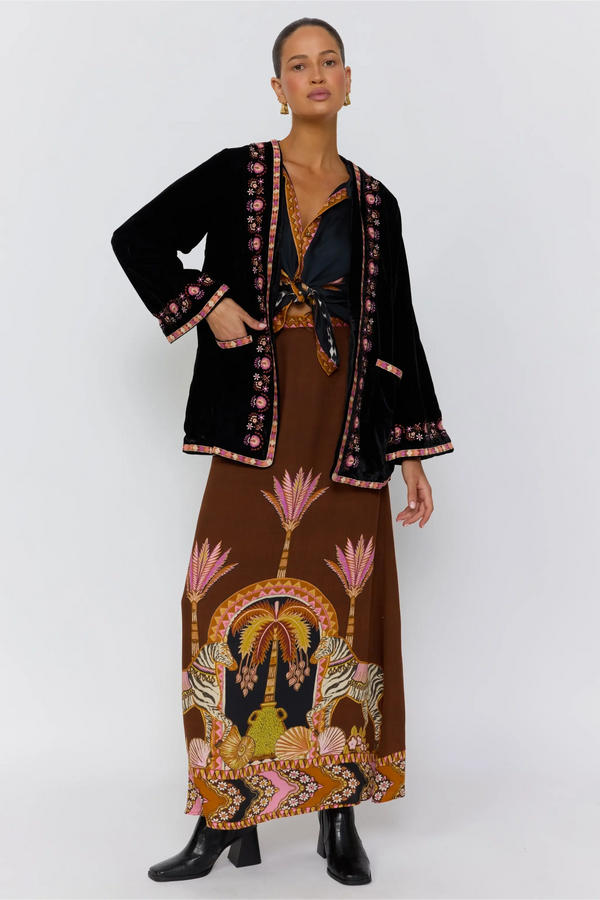 Woman wearing a black jacket with floral patterns and a long brown dress with a decorative design on a white background