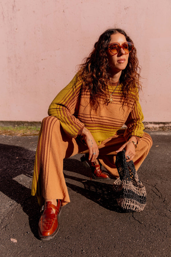 Woman in mustard sweater and beige pants sitting on a curb with a pink wall background