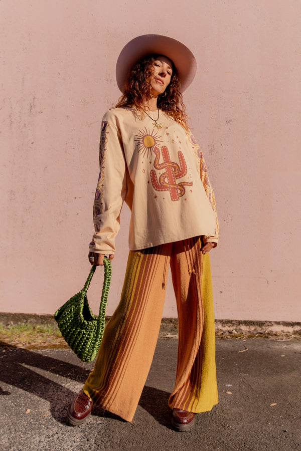 Woman wearing a beige sweatshirt with a graphic design, brown hat, and colorful pants against a pink wall.