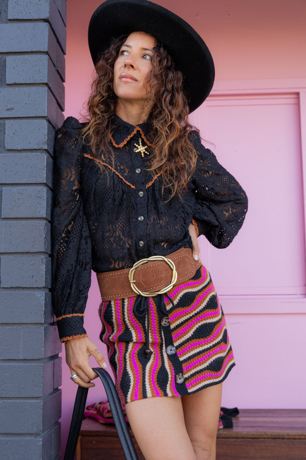 Woman in a stylish outfit with a black hat, black lace top, and colorful skirt against a pink wall.