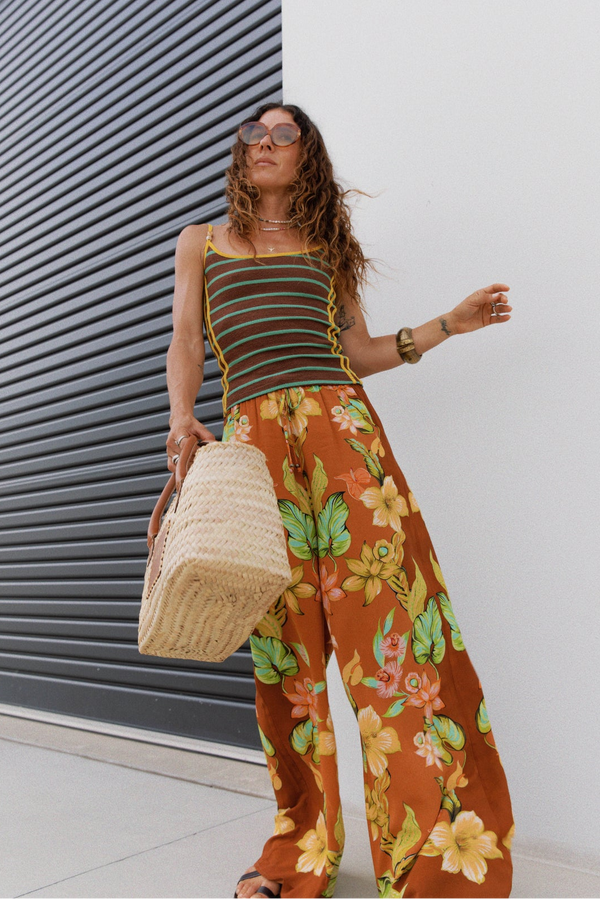 A woman wearing a brown and orange striped camisole top and matching wide-leg floral pants in rust orange, holding a woven straw tote bag, posing in front of a white wall and a dark gray metal shutter.