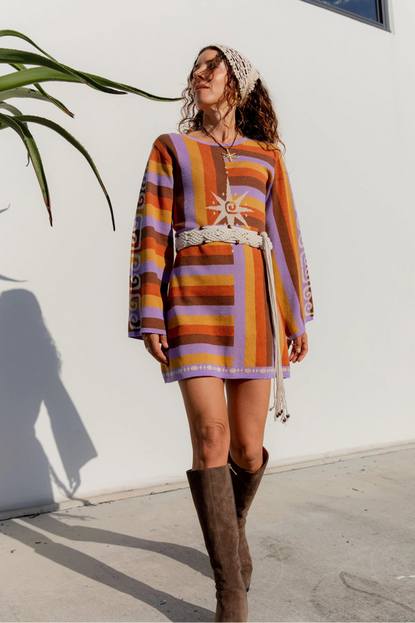 Woman in a colorful striped dress and brown boots standing against a white wall.