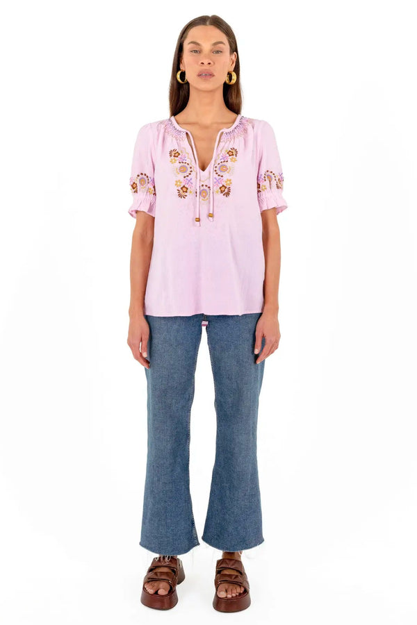 A woman modeling a light pink short-sleeve blouse with colorful floral embroidery on the chest and sleeves, paired with blue flared jeans and brown sandals, against a plain white background.