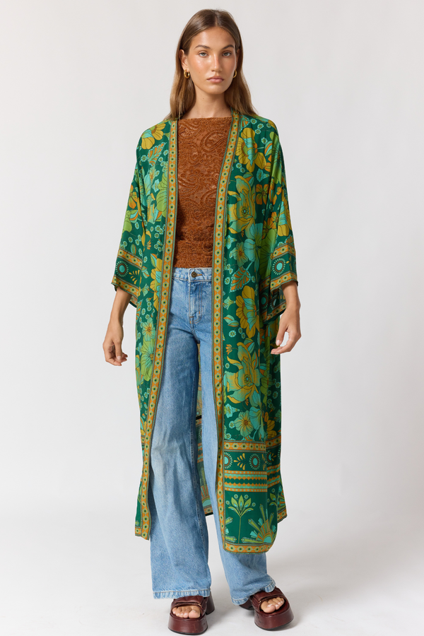 Woman wearing a long green patterned robe over a brown sweater and blue jeans on a white background