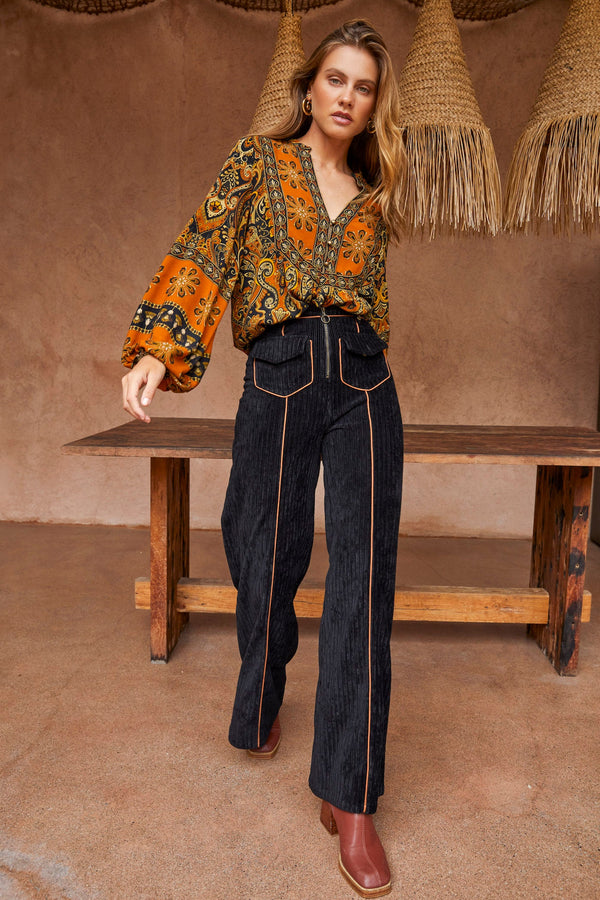 Woman in a patterned blouse and jeans standing in a rustic setting with wooden table and straw hats.